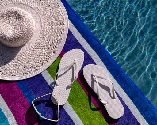 Pair of white flip flops by the pool on a bright blue, green, purple and white striped towel with sunglasses and big white floppy hat